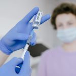 Profissional de saúde com luvas azuis prepara uma seringa com vacina, enquanto um paciente aparece desfocado ao fundo.