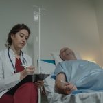 Médica consulta um prontuário ao lado de um paciente em um quarto hospitalar.