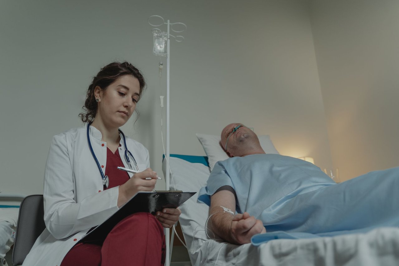 Médica consulta um prontuário ao lado de um paciente em um quarto hospitalar.