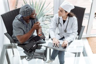 Dois médicos discutem cuidados de saúde usando um laptop e um tablet em ambiente clínico.