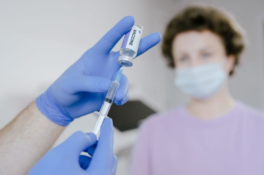 Profissional de saúde com luvas azuis prepara uma seringa com vacina, enquanto um paciente aparece desfocado ao fundo.